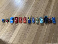 Wide shot of all 13 Disney Pixar Cars diecast toy vehicles arranged in a row on a wooden floor, showing size and color variety.