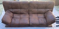 Brown faux leather futon couch with visible tufting and wide armrests on outdoor surface.