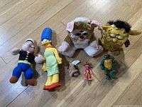 Group photo of 8 plush toys and action figures on wooden floor including 2 Furbies, Popeye, Marge Simpson, Pink Panther, TMNT, and small brown animal figure