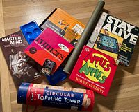 Photo showing six boxed games and a cylindrical game container arranged on wood floor including Deluxe Master Mind, Waddingtons Basket Ball, Timors card game, Apples to Apples Jewish Edition, Stay Alive, and Circular Toppling Tower.