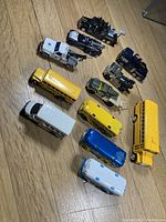 Top view of twelve diecast toy vehicles on wooden floor including tow trucks and buses in various colors and sizes.