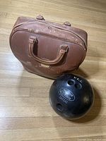 Black Manhattan Rubber bowling ball and vintage brown Brunswick bowling ball bag placed on wooden floor; ball shows three finger holes and marking details; bag shows wear and scuffs.
