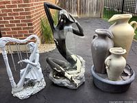 Outdoor photo showing three garden fountains and sculptures including woman silhouette, water jug fountain, and lady playing harp.