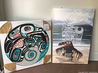 Two Native art canvases leaning against wall, one circular signed with black, turquoise, red and white elements and the other rectangular with a black bird figure on a light background with white and gray shapes