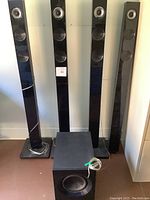 Front view of 4 black LG tower speakers and 1 square black subwoofer speaker on floor. One tower speaker is missing its base.