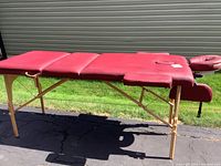 Full side angle view of maroon padded massage table with wooden legs on asphalt, outdoor lighting.