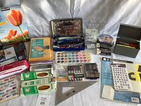 Wide view of the assortment of office and art supplies displayed on a white surface, showing paper products, pens, watercolor palette, staples, and stickers.