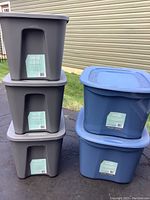 Stack of three gray 40L Restore Totes and stack of two blue 63L Prima Storage Containers on driveway.