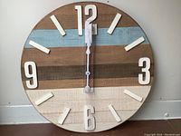 Front view of large round wooden farmhouse clock with white 3D numbers and hour markers on a multi-colored horizontal wood plank background.