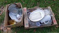 Two boxes filled with assorted vintage glassware and dinnerware including glass bowls, plates, cups, and containers of various sizes and ages, showing overall lot contents on grass background.