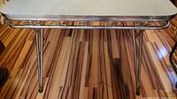 Full view of mid-century dining table with stainless steel legs and edge trim, standing on wood floor.