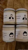Four glass jar candles with white lids arranged closely showing labels with illustrations and quotes from Schitts Creek characters David and Johnny.