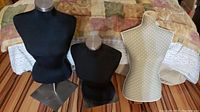 Three small dress forms on a bed, two black covered and one beige with polka dots, metal bases visible.