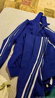 Blue vintage track suit laid on surface showing folded jacket and pants, visible white stripes on sleeves and legs.