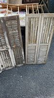Three vintage wooden window shutters placed on asphalt, showing varying degrees of weathering and chipped paint.