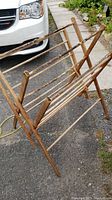Side angled view showing the wooden clothes rack positioned on an asphalt surface beside a white vehicle and garden area.