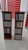 Two vertical wooden CD towers filled with assorted CDs, positioned side by side against a red storage unit background.