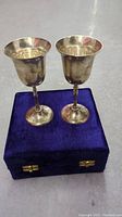Two silver plated goblets resting on top of a closed purple velvet case with gold-colored metal clasps.
