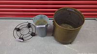 Wide view showing all three items together against a red door backdrop: an antique bronze planter, metal plant stand with wheels, and galvanized steel bucket planter.