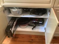 Photo showing suite of fry pans stacked inside cabinet with salad spinner on upper shelf and Fox Run hot stone set box on lower shelf.