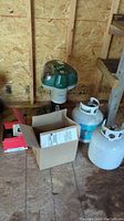 Green and white Mosquito Magnet device with two white propane cylinders and boxes on floor in shed.