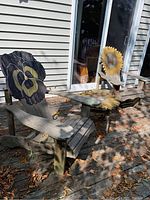 Pair of two wooden Muskoka chairs with flower designs on backs and a wooden table between them on outdoor wooden deck with fallen leaves and siding background