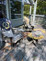 Photo showing two weathered wooden Muskoka chairs with hand painted floral designs on the backrests and a matching painted low rectangular table on a wood deck outside.