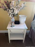 White night stand with a vase of faux flowers, woven basket, alarm clock, and figurine on top