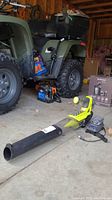 Whole lot showing the Ryobi cordless leaf blower, batteries, and charger placed on concrete floor near a green ATV.