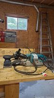 Three power tools on a wooden table in workshop: Makita belt sander, Mastercraft 1/4 sheet sander, Jobmate angle grinder.