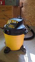 Front angle of Dirt Hawk 12 gallon 5.0 HP wet-dry vacuum, yellow tank, black motor top, attached hose with duct tape.