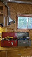 Red Craftsman and grey Mastercraft metal toolboxes on a wooden countertop under a window, both open showing top trays filled with hand tools like screwdrivers and pliers.