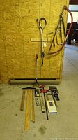 Photo showing multiple measuring sticks of various lengths and materials, a large bow saw with an orange handle, a long snipped metal tool, and a pair of bolt cutters hanging on the wall.
