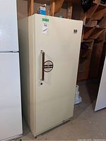 Front view of the upright freezer showing cream color, vertical handle, and overall cupboard-style door.