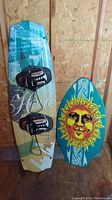 Photo showing both the 55-inch Hobie wakeboard with bindings and the 36-inch skim board with sun face design leaning against plywood wall.