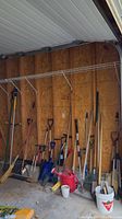 Wide view of assorted yard and garden tools arranged against a plywood wall in a garage or shed area, showing various long-handled tools like shovels, rakes, and trimmers.