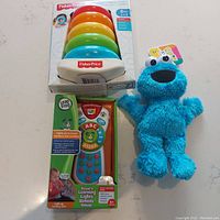 Photo of all three toys including the Fisher-Price Rock a Stack pyramid in packaging, the LeapFrog Scout's Learning Lights Remote Deluxe box, and the plush Cookie Monster toy. Shows condition and packaging of items.