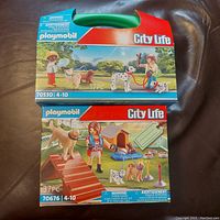 Front view of Playmobil City Life 70530 and 70676 sealed boxes with green handles and packaging artwork showing figures with dogs and accessories.