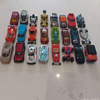 Top-down photo showing all 24 Hot Wheels cars lined up in four rows on a white surface. Variety of colors and shapes visible including sports cars, race cars, trucks, and futuristic cars.