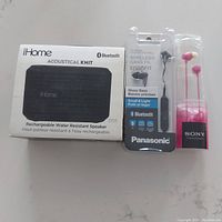 Photo showing the iHome Acoustical Knit Bluetooth speaker in packaging next to Panasonic wireless Bluetooth earbuds and Sony pink corded earbuds in original sealed packaging.