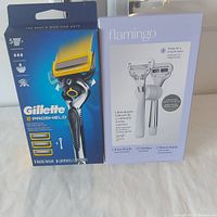 Front view of Men's Gillette Proshield razor package with 3 cartridges and Ladies Flamingo razor package with 1 handle, 2 cartridges, and shower holder shown side by side.