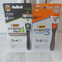 Photo of two sealed boxes of Bic men's razor products. Left box is Bic Hybrid Flex 4 Titanium Sensitive with 1 handle and 4 cartridges. Right box is Bic Hybrid Comfort 3 with 1 handle and 6 cartridges.