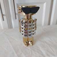 Front side of gold-colored Paco Rabanne Fame perfume bottle shaped like stylized figure with shiny scale pattern and black butterfly mask.