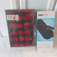 Full view of Aroma Homme hot water bottle in red and black plaid cover alongside Obusforme supporting roll in box.