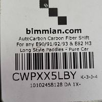 Close-up photo of label from bimmian.com showing product details: AutoCarbon Carbon Fiber Shift for BMW E90/91/92/93 & E82 M3 Long Style Paddles - Pure Carbon.