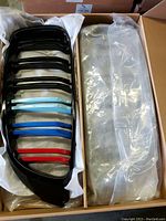 Open box showing a gloss black kidney grille with colored dual slats and protective plastic wrap inside cardboard packaging