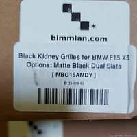 Close-up photo of a label on one box showing the product as Black Kidney Grilles for BMW F15 X5 with Matte Black Dual Slats option by bimmian.com, product code MBG15AMDY.