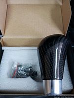 Close-up of a carbon fiber gear shift knob with silver base and installation hardware packaged in white foam inside an open box.