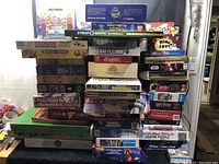Stacked assortment of board games and puzzles piled high on a table, showing variety of titles and box sizes.