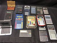 Lot of 15 assorted calculators on black fabric background including handheld and pocket styles, various colors and button configurations.